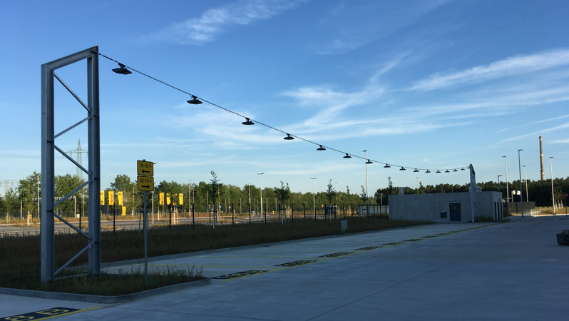 Industriegelände mit Lampen an einem Drahtseil, Zäunen und Schildern unter blauem Himmel. Referenz Elektrotechnik Rudolf Fritz GmbH