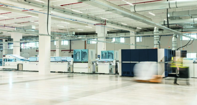 Moderne, helle Fabrikhalle mit industriellen Maschinen und Produktionsanlagen. Im Vordergrund bewegt sich eine unscharfe Person in Sicherheitskleidung mit einer Palette. Referenz Rudolf Fritz GmbH