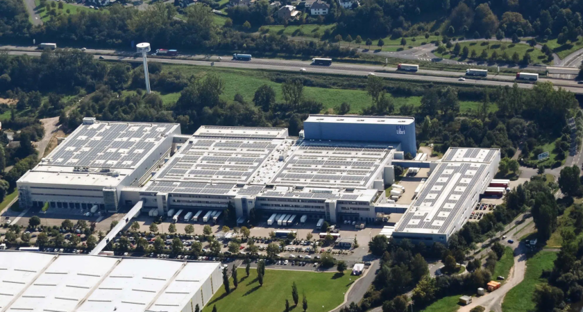 Luftaufnahme eines großen Industrie- oder Logistikkomplexes mit mehreren flachen Gebäuden, deren Dächer mit Solarpanelen bedeckt sind. Referenz Rudolf Fritz GmbH